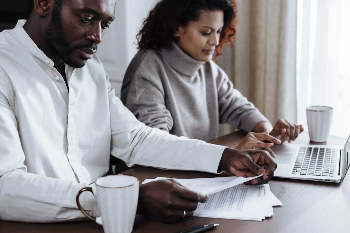 A man and a woman sit at a table reviewing documents beside a laptop. A man and a woman sit at a table reviewing documents beside a laptop.