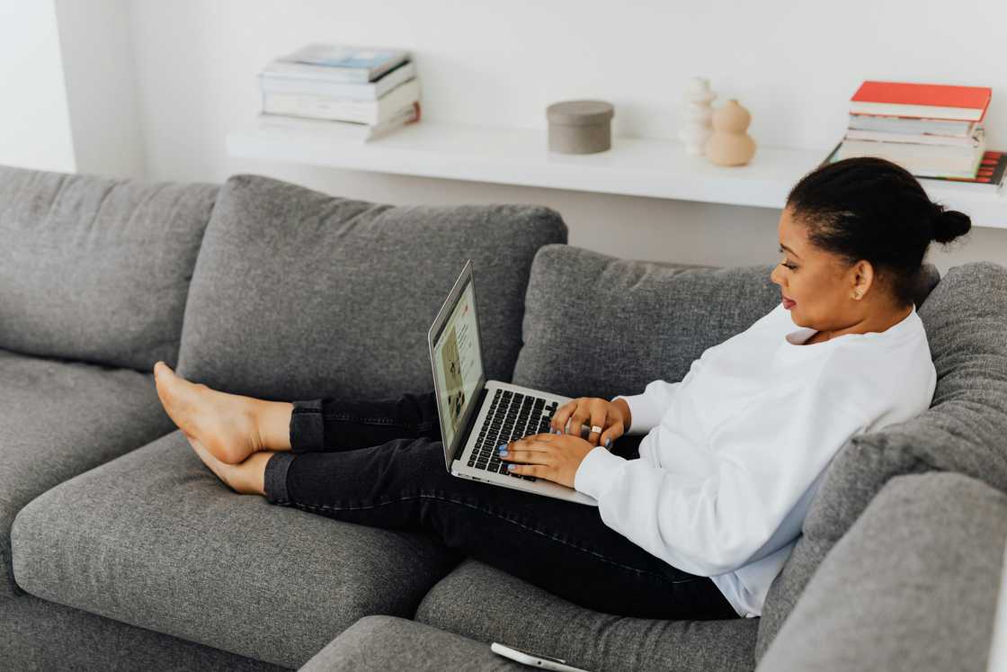 A woman in a white top and black pants is using a laptop on a grey couch