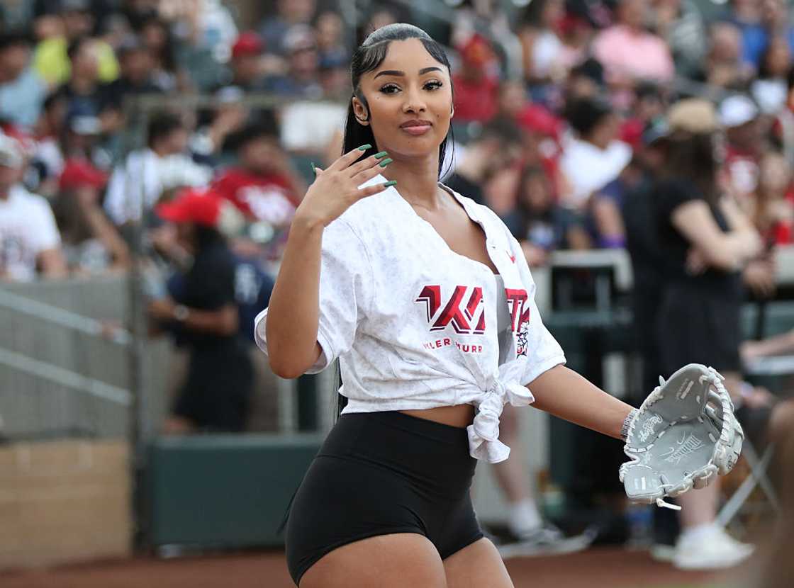A woman in a tied white jersey and black shorts waves while wearing a baseball glove on the field.