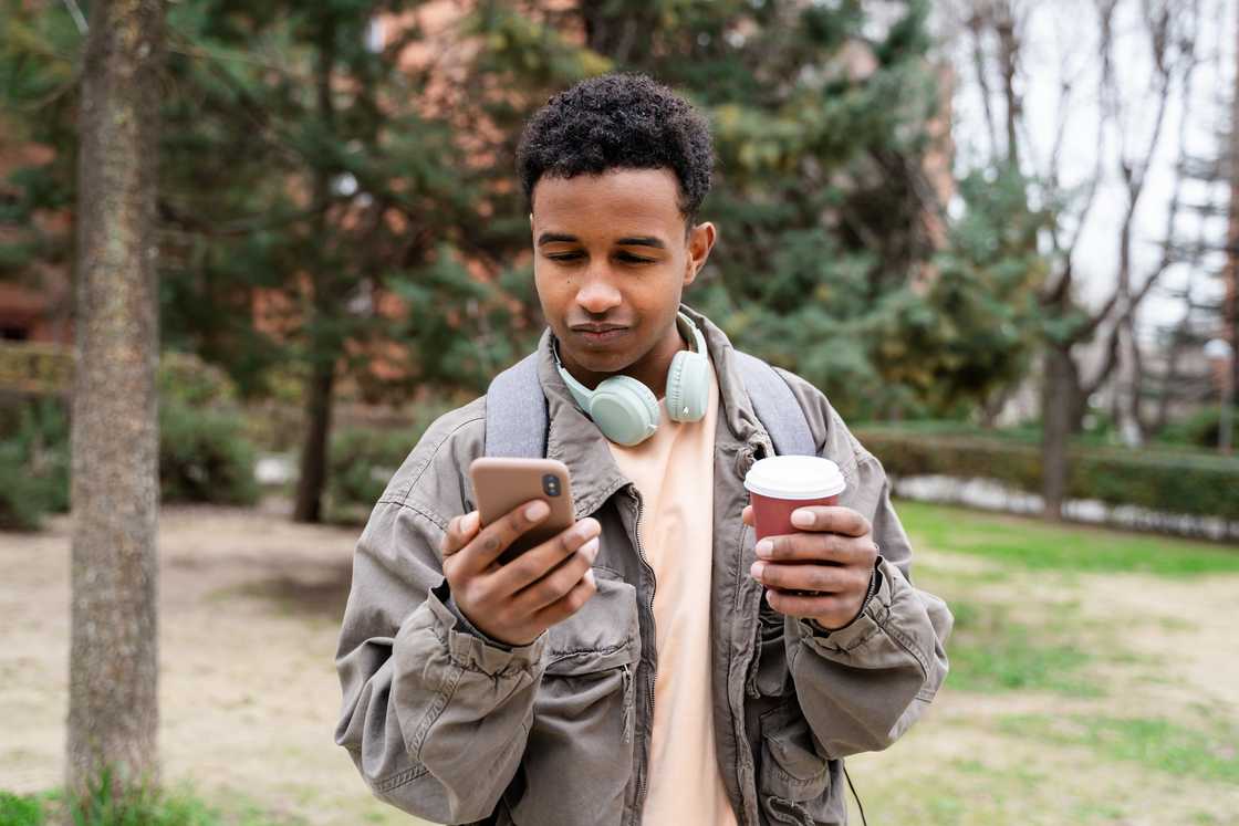 Young man using smartphone and drinking coffee in an urban park setting.