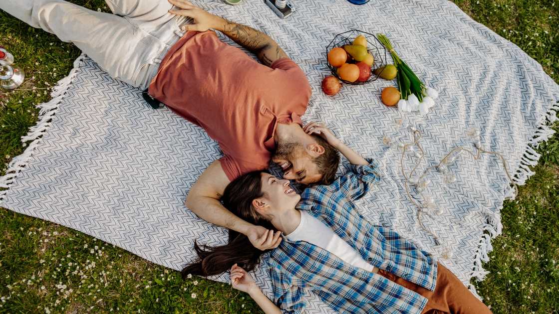 A man in an orange shirt and a woman in a patterned blue shirt look at each other while lying on a grey blanket on the ground.