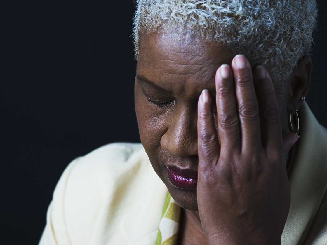 Person with short gray hair covers one eye with a hand, wearing a light blazer against a dark background. Person with short gray hair covers one eye with a hand, wearing a light blazer against a dark background.