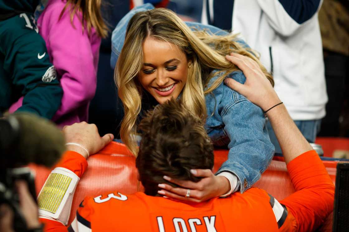 A blonde woman in a denim jacket hugs a football player wearing a "Lock" jersey from above. A blonde woman in a denim jacket hugs a football player wearing a "Lock" jersey from above.