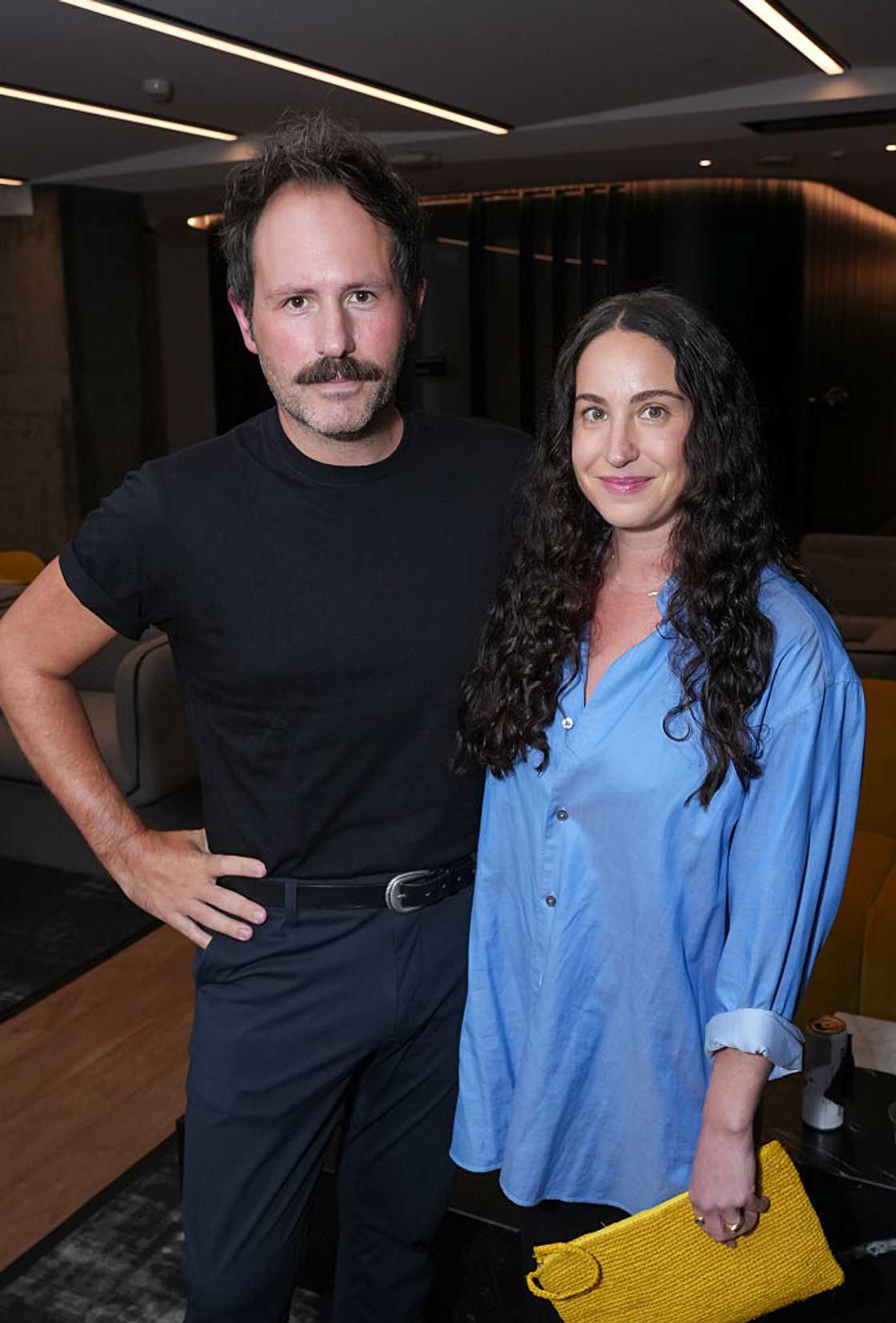 A mustached man in a black T-shirt stands beside a curly-haired woman in a blue blouse holding a yellow clutch. A mustached man in a black T-shirt stands beside a curly-haired woman in a blue blouse holding a yellow clutch.