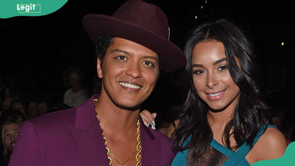 Bruno Mars (L) and Jessica Caban pose together at a nighttime event with a crowd blurred in the background