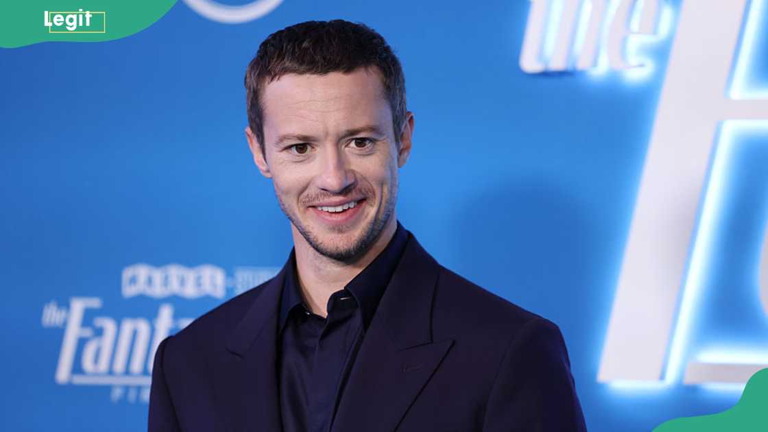 A man in a dark navy suit and shirt smiles in front of a blue Fantastic Four premiere backdrop. A man in a dark navy suit and shirt smiles in front of a blue Fantastic Four premiere backdrop.