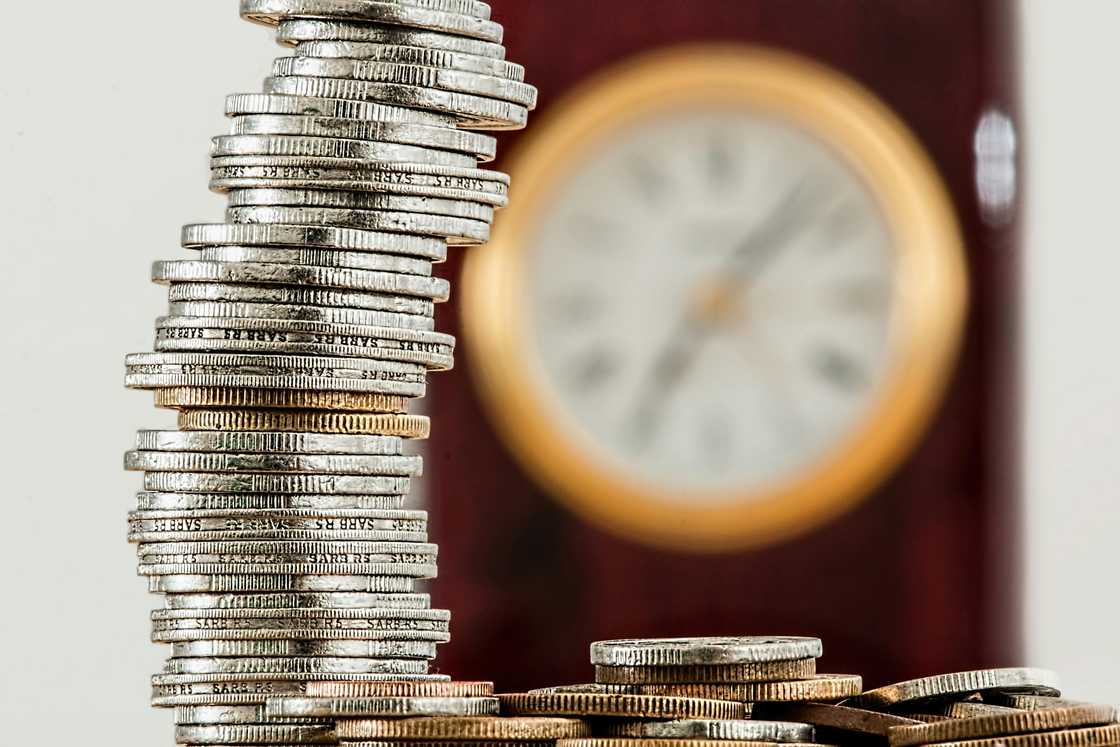 Coins stacked on top of each other in front of a yellow wall clock Coins stacked on top of each other in front of a yellow wall clock