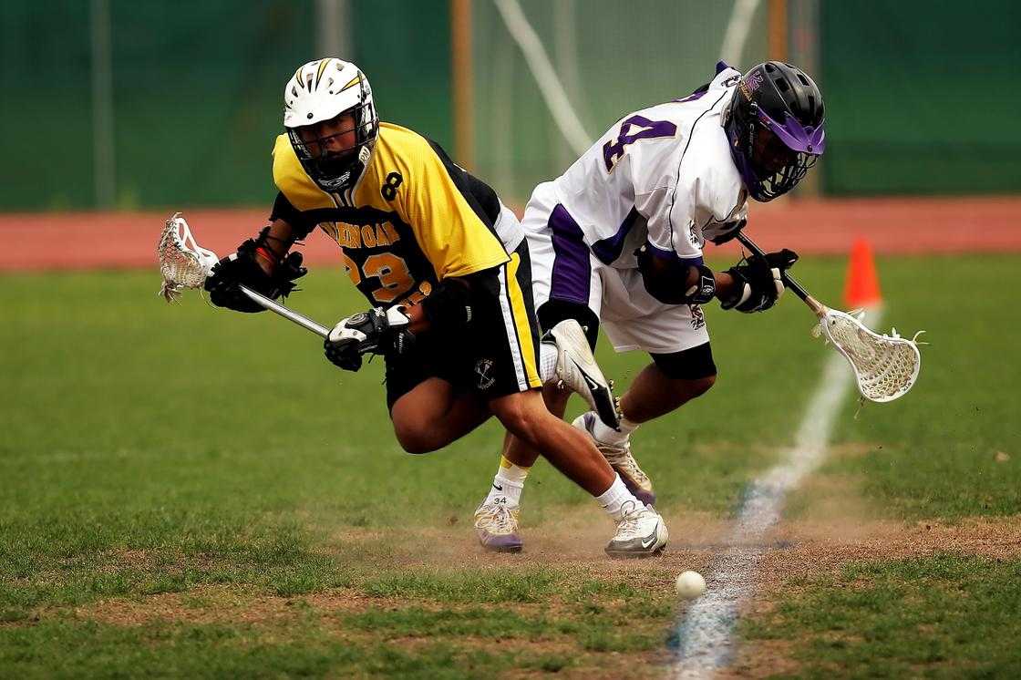 A Man wearing a yellow and a black sport jersey holding lacrosse sticks.