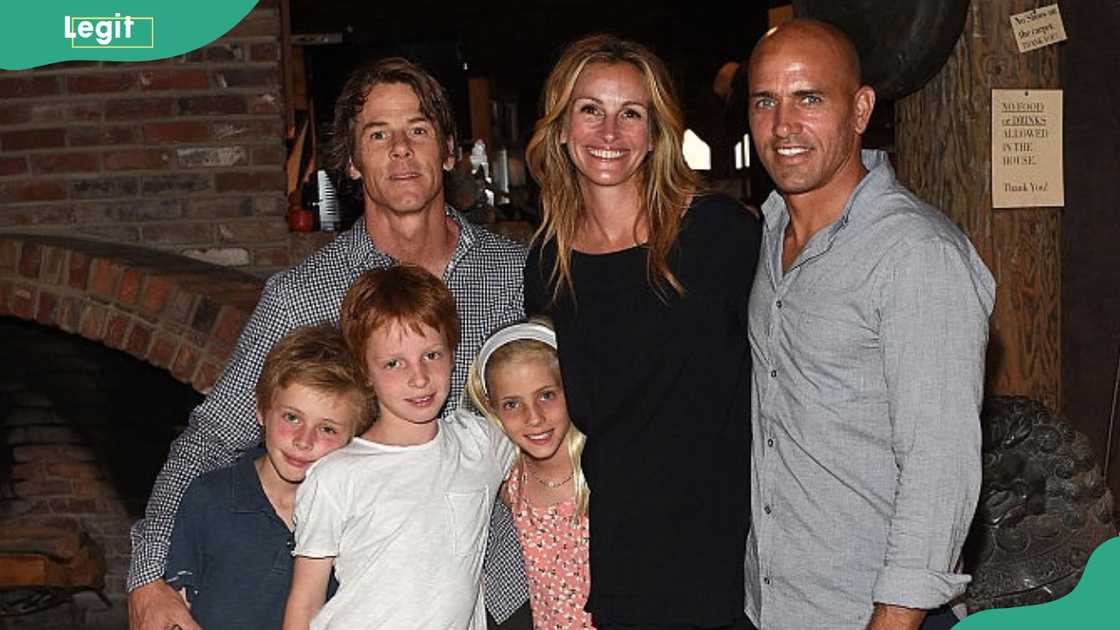 A woman in a black dress stands with a man, three children, and another man in a grey shirt indoors. A woman in a black dress stands with a man, three children, and another man in a grey shirt indoors.
