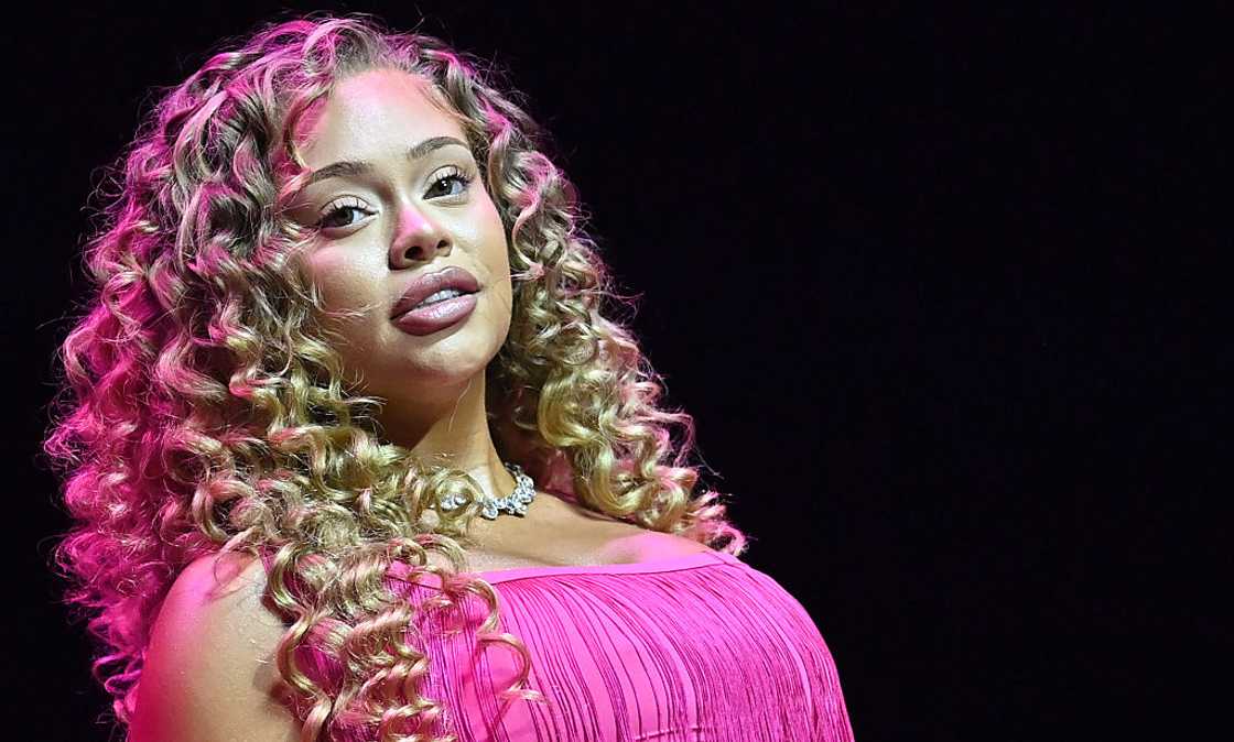 Rapper Latto with long curly hair poses on stage in a pink off-shoulder gown lit with pink light.