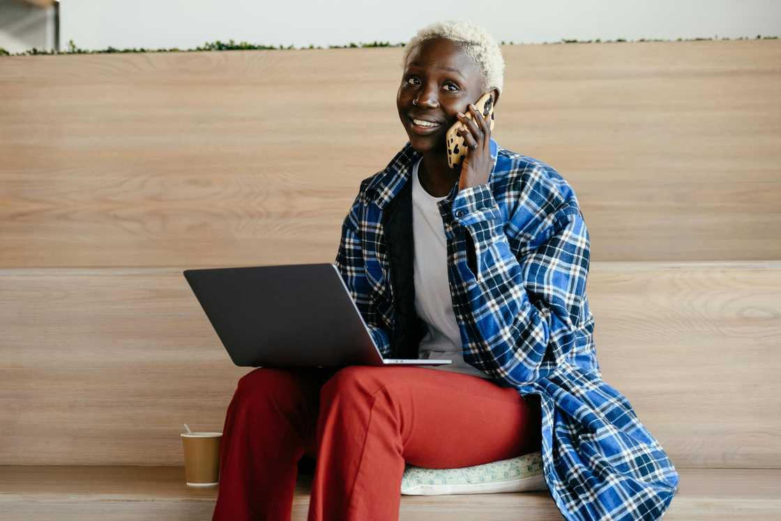 A young woman with blonde hair, a blue checkered shirt, and maroon pants is using a laptop as she talks on the phone