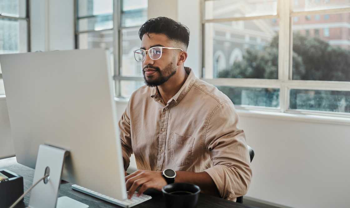 A young man in a beige sweater works on a desktop computer in a bright modern office, representing an online job application. A young man in a beige sweater works on a desktop computer in a bright modern office, representing an online job application.