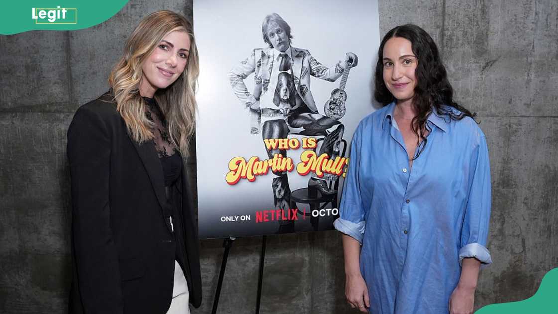 A blonde woman in a black blazer and a dark-haired woman in a blue shirt stand beside a film poster on an easel.