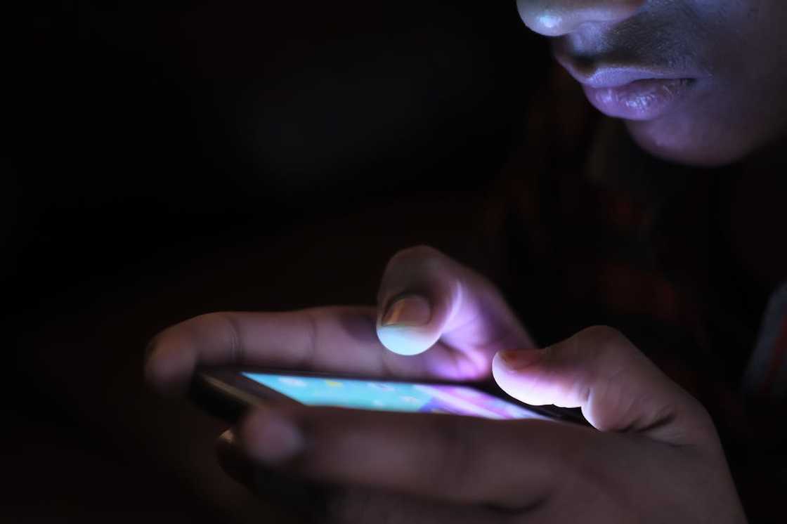 A person scrolls on a smartphone in the dark, lit by the screen.