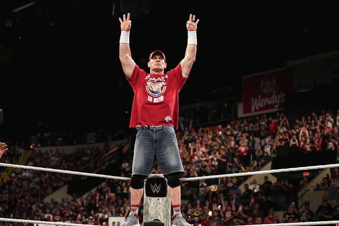 John Cena in a red T-shirt and jean shorts raising both arms to the crowd from the ring at the MassMutual Center in Springfield, Massachusetts, 2025