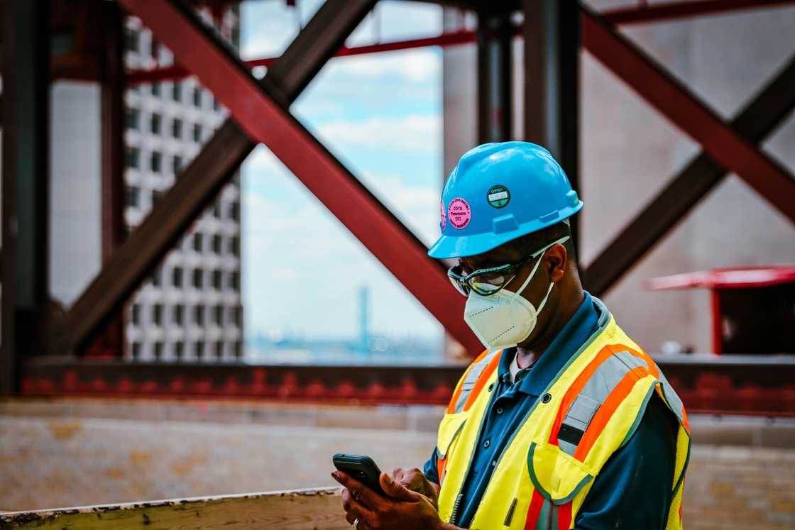 An engineer wearing a hat, mask, and reflector jacket checking his phone An engineer wearing a hat, mask, and reflector jacket checking his phone
