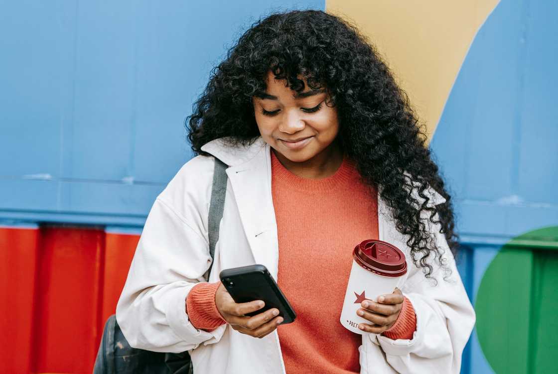 A woman using her smartphone standing near a colour wall A woman using her smartphone standing near a colour wall