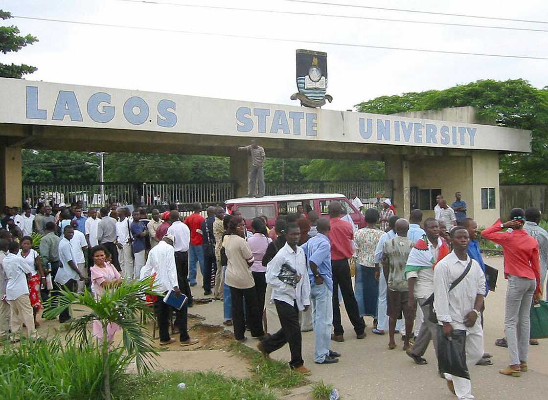 A large group of people stand and walk near the Lagos State University entrance archway, with a van and greenery in the background.