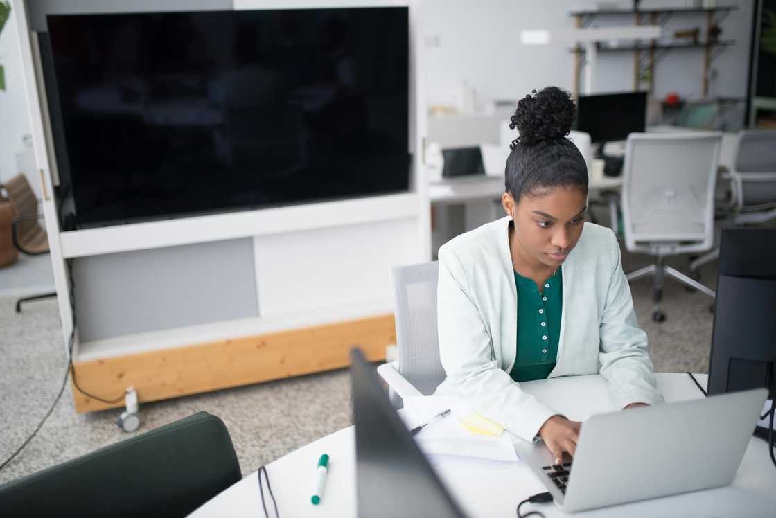 A woman in a green top and white blazer is using a laptop on the desk