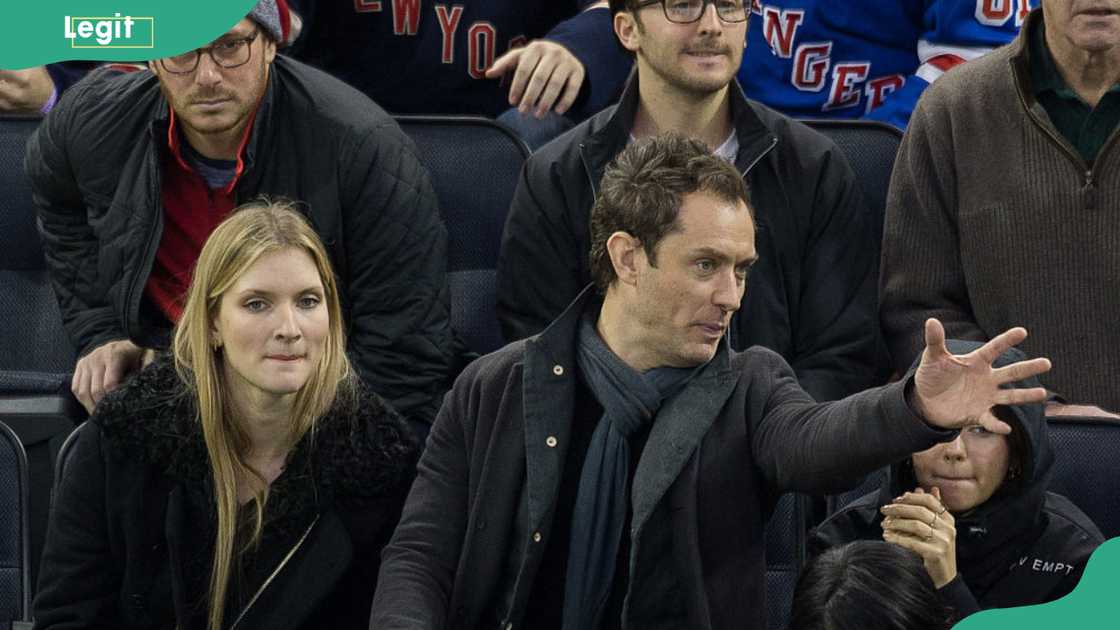Jude Law gestures with his hand while seated among fans at a Rangers game.