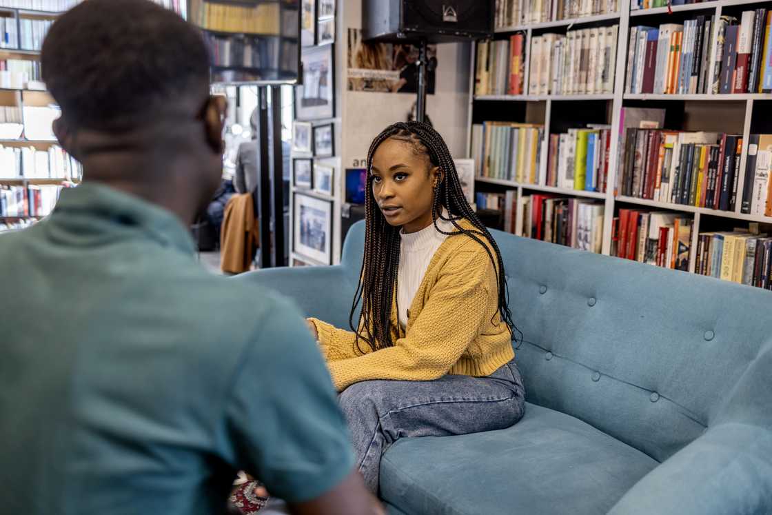 Two people talk on a couch in a room lined with bookshelves. Two people talk on a couch in a room lined with bookshelves.