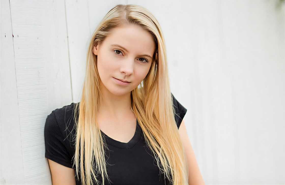 Alison Bassett, poses in a black v-neck top, leaning against a white textured wall. Alison Bassett, poses in a black v-neck top, leaning against a white textured wall.