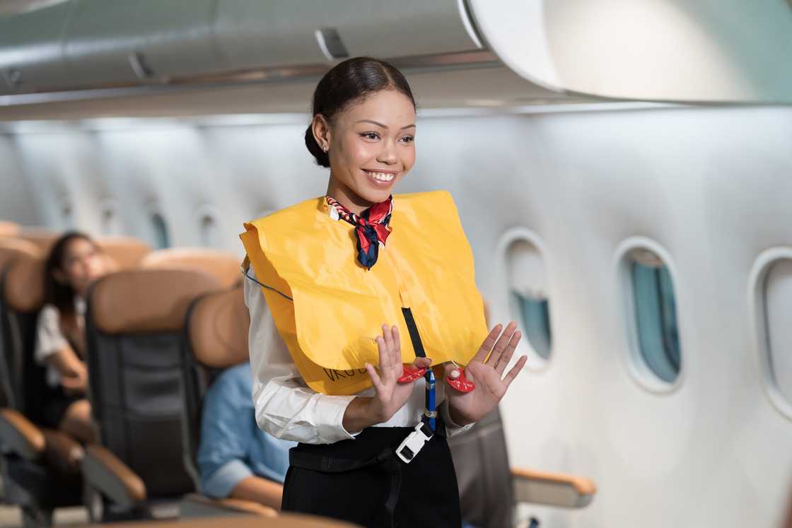 A smiling flight attendant in a yellow life vest holds up the vest's red toggles in an aeroplane cabin.