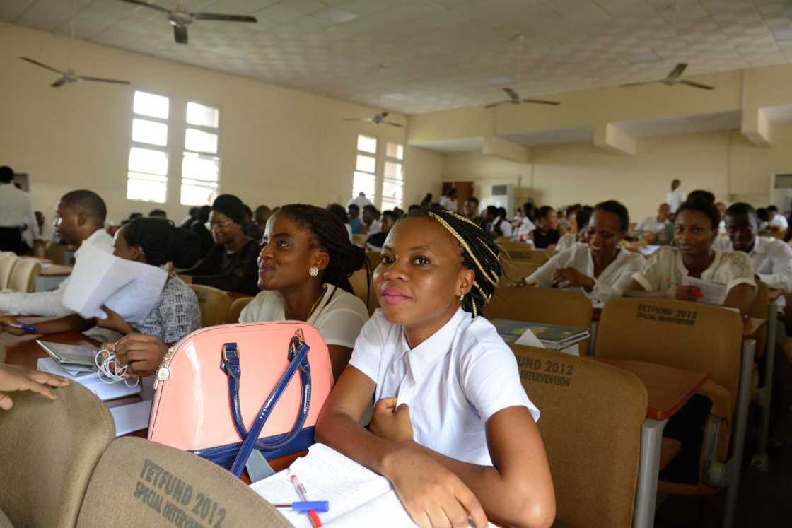 A young woman with braided hair sits at a desk beside a pink handbag, surrounded by other students seated in rows under ceiling fans. A young woman with braided hair sits at a desk beside a pink handbag, surrounded by other students seated in rows under ceiling fans.