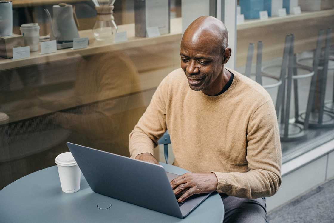 A mature black man in a brown sweater and grey pants is using a laptop as he takes coffee