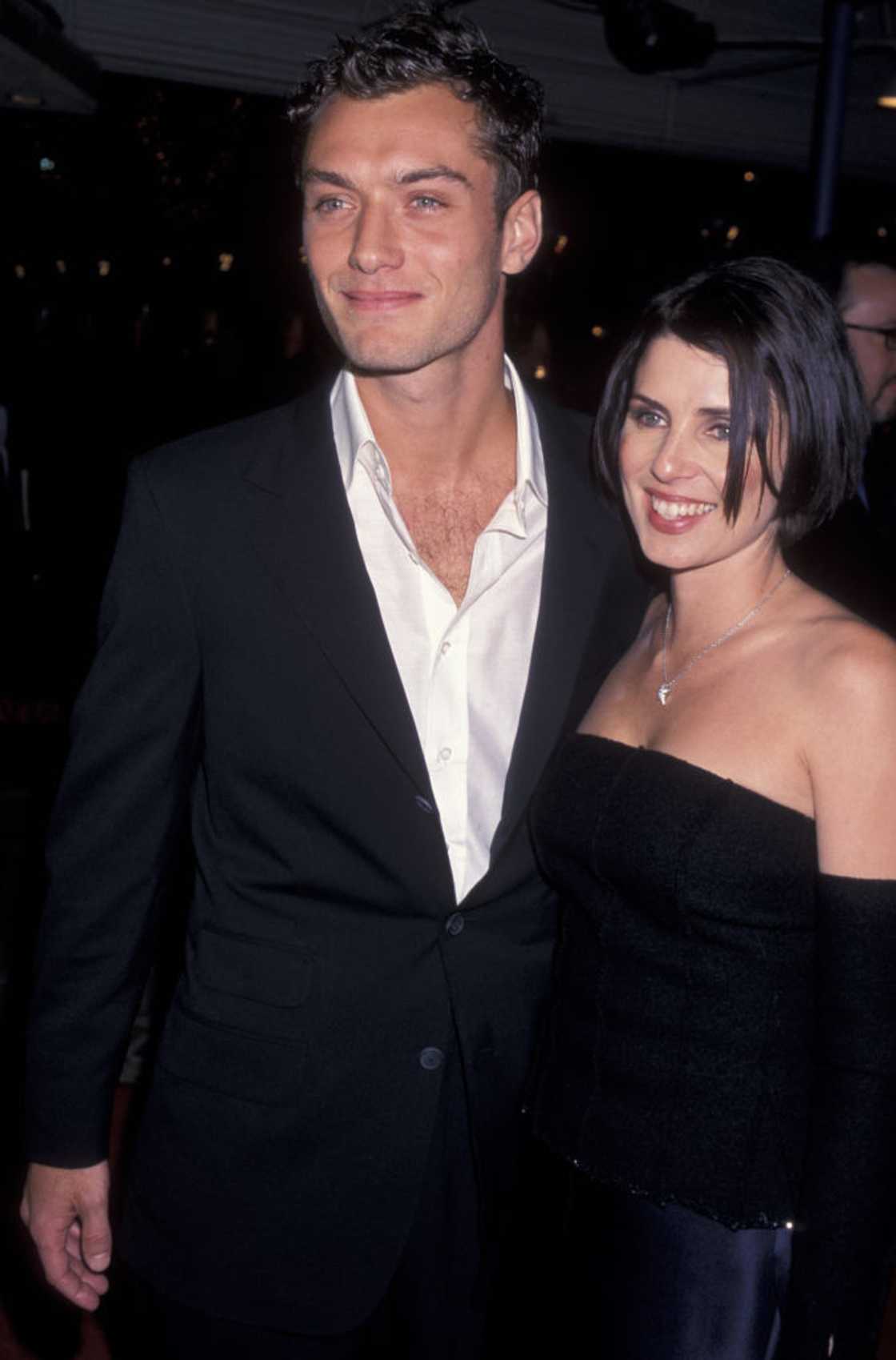 A younger Jude Law in a black suit stands beside a woman in a black strapless top with a necklace. A younger Jude Law in a black suit stands beside a woman in a black strapless top with a necklace.
