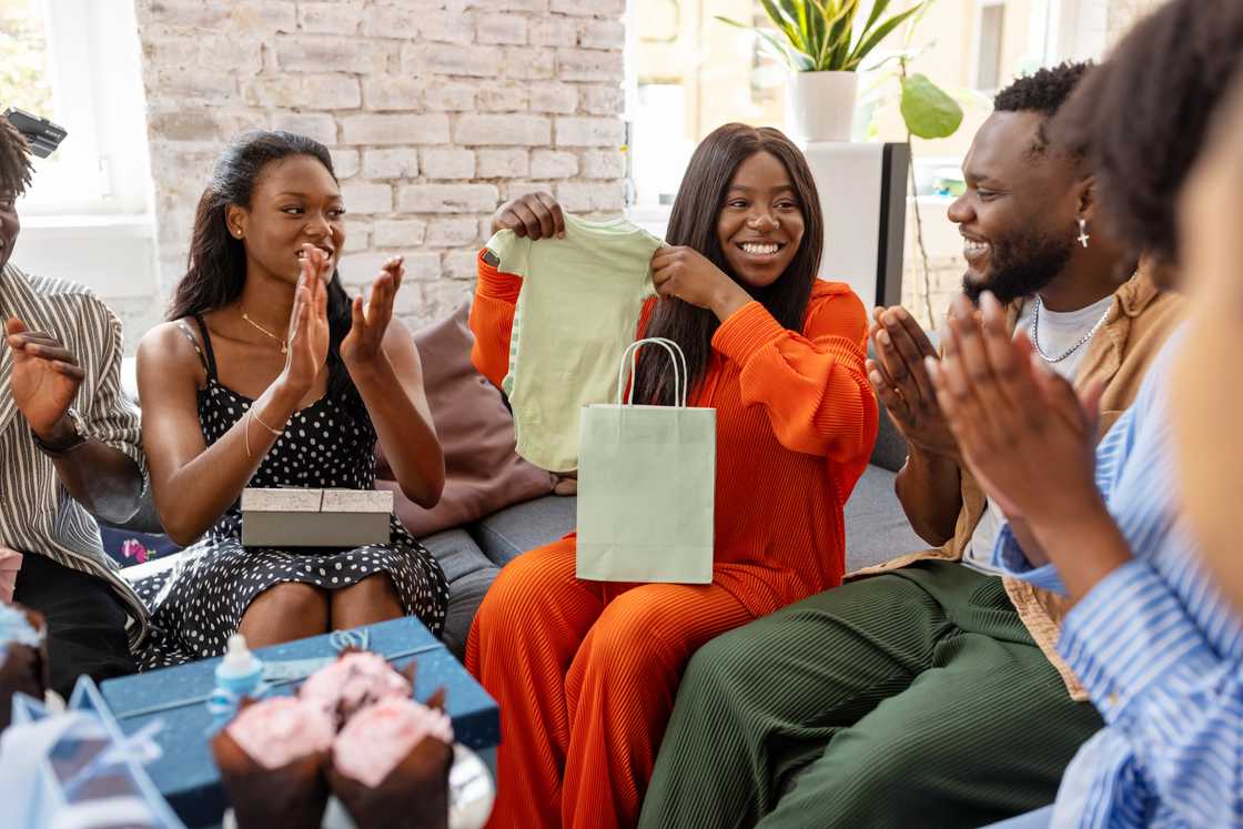 A young pregnant woman getting baby clothes as a present from her friends at a baby shower party.