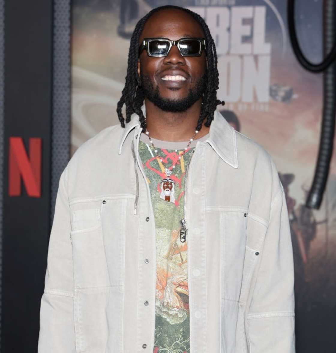 Dot da Genius in sunglasses and a cream jacket poses in front of a "Rebel Moon" Netflix backdrop.