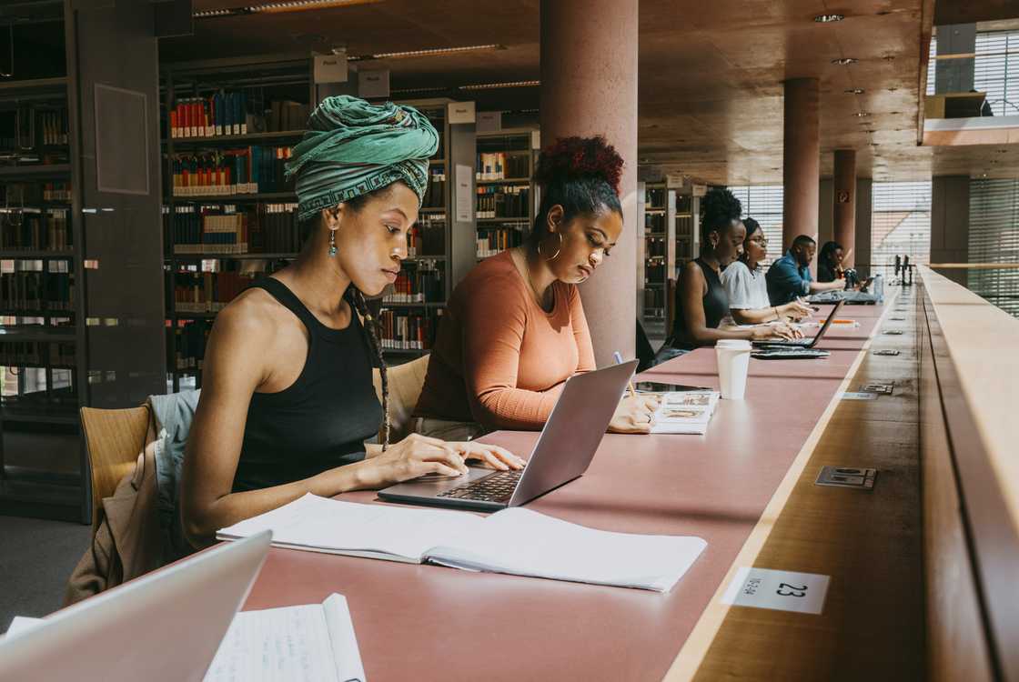 A row of students, including one wearing a green head wrap, sit at a library table typing on laptops and taking notes. A row of students, including one wearing a green head wrap, sit at a library table typing on laptops and taking notes.