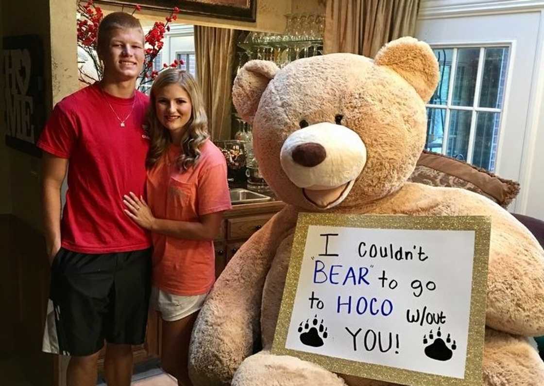 A couple posing for a photo next to a giant teddy bear with a Hoco proposal