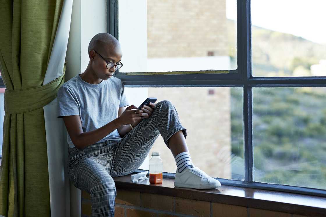 A lady sitting on a window sill with juice while scrolling her phone