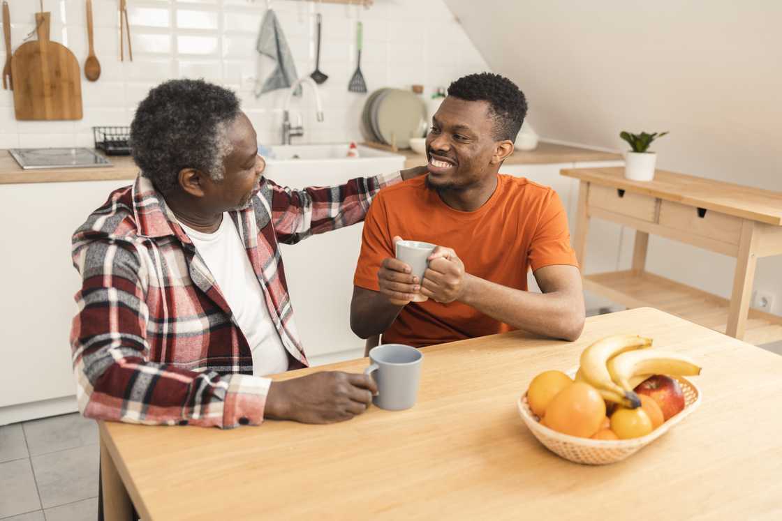 Two people sit at a kitchen table with mugs and a bowl of fruit.
