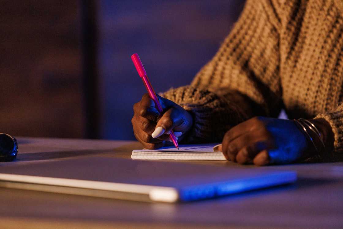 Person in brown sweater writes in a notebook with a red pen beside a closed laptop.