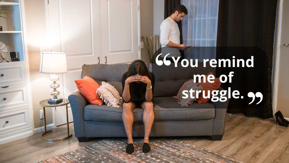 A woman sits on a sofa with her head in her hands while a man stands behind her checking his phone.