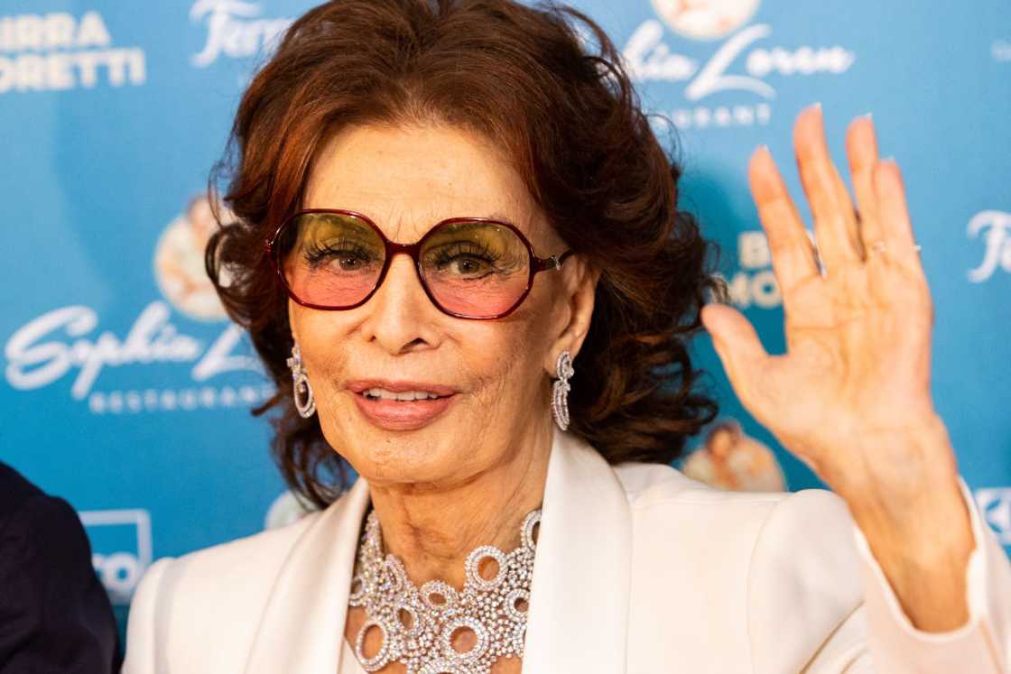 Italian actress Sophia Loren waves in sunglasses, a pearl necklace and a white outfit seen at Sophia Loren Restaurant in Milan