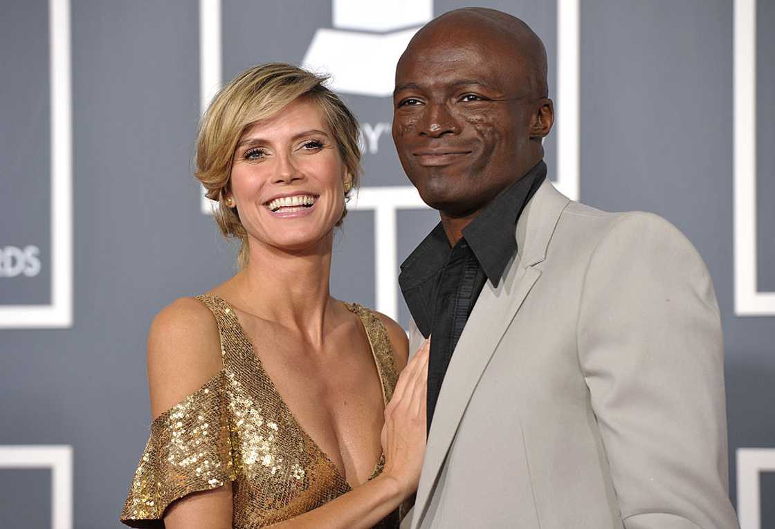 A bald man in a light beige jacket over a black shirt stands close to a woman with short blonde hair. A bald man in a light beige jacket over a black shirt stands close to a woman with short blonde hair.