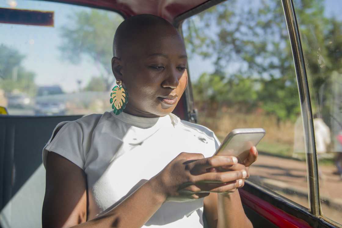 A woman with a shaved head and green leaf-shaped earrings looks down at her phone, sunlight streaming through the vehicle window behind her.