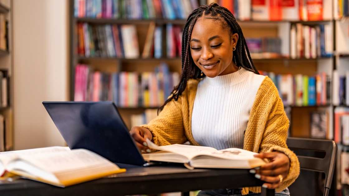 A female student reads a book in front of a laptop in a library.