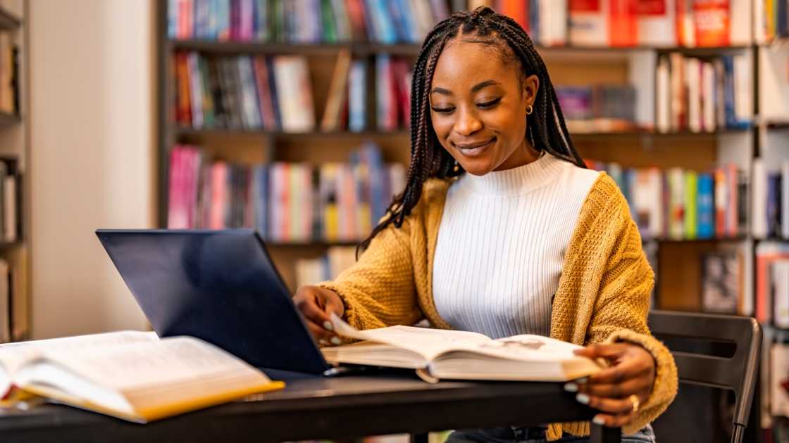 A female student reads a book in front of a laptop in a library. A female student reads a book in front of a laptop in a library.