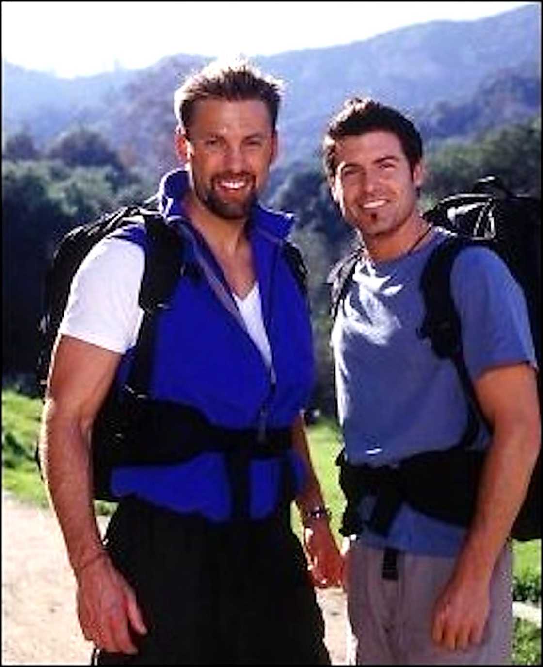 A man in a blue life vest with a backpack stands beside another man in a grey shirt with a backpack, hills and trees behind them.
