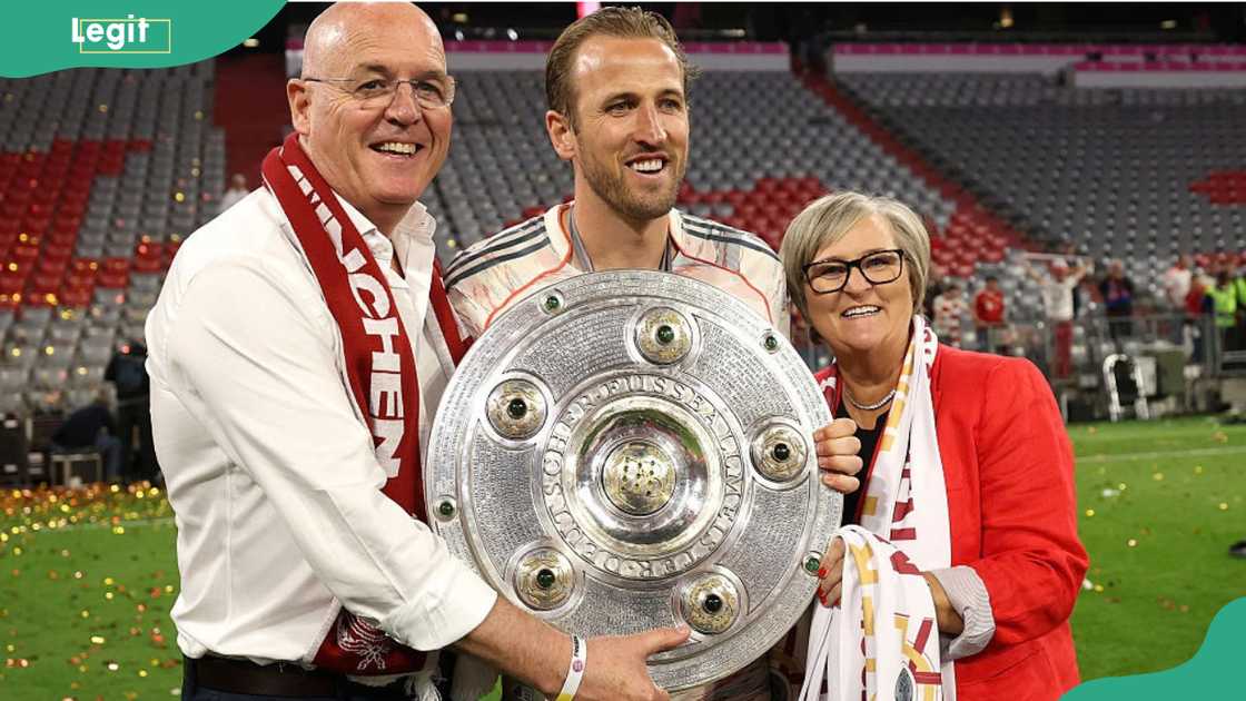 A man in a football kit holds a large silver trophy between an older man and woman in red scarves. A man in a football kit holds a large silver trophy between an older man and woman in red scarves.