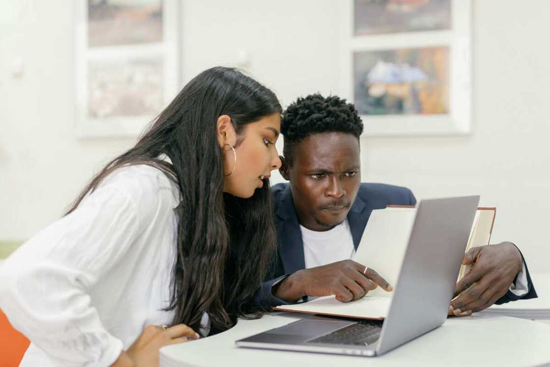 A professor and a student sitting at a desk with a computer and a book A professor and a student sitting at a desk with a computer and a book