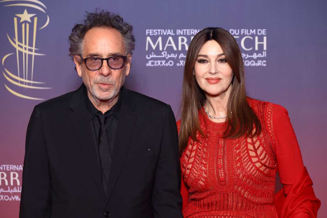 A man in a black suit stands beside a woman in a red beaded gown at a festival backdrop.