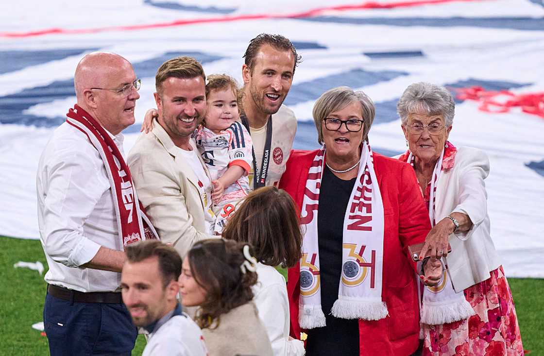 A group including two men, a child, and two older women in red scarves smile together on a football pitch. A group including two men, a child, and two older women in red scarves smile together on a football pitch.