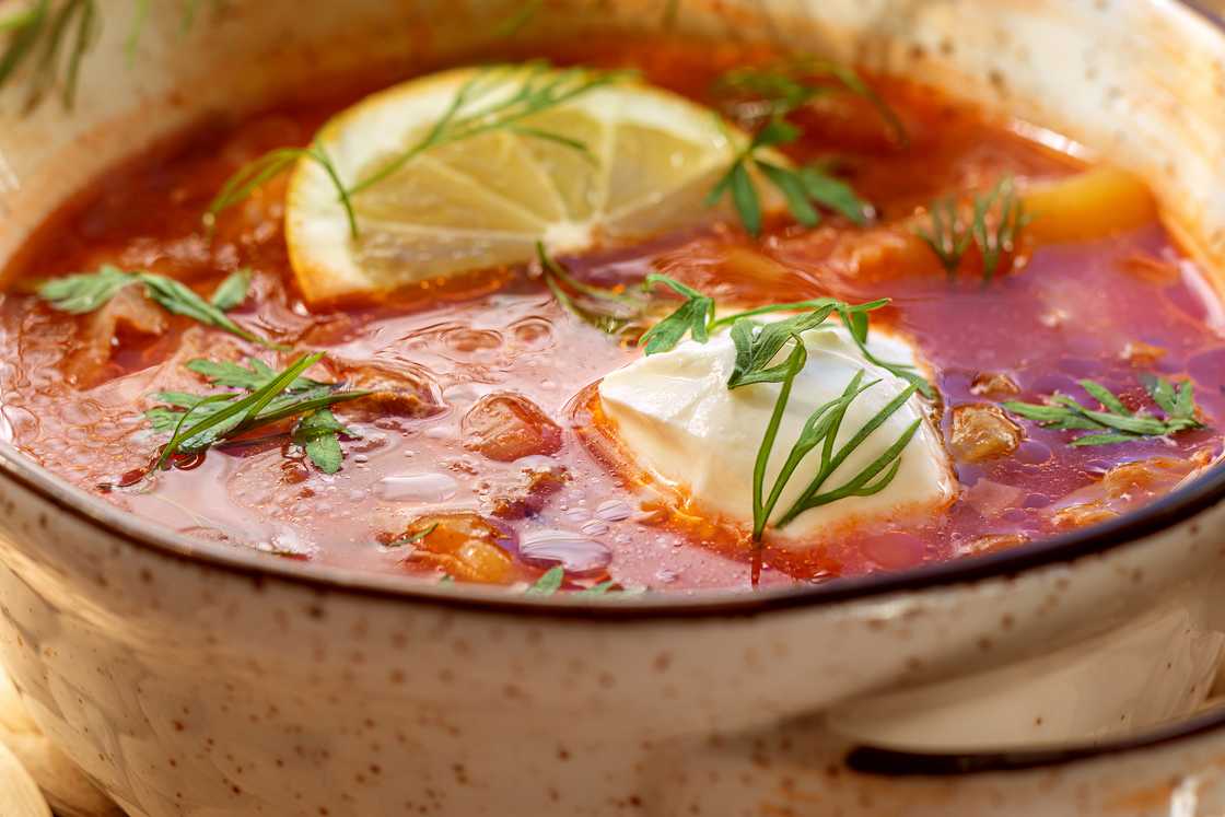 Vegetable soup in saucepan. Bright red soup with fresh cabbage. Decorated with lemon slices and chopped dill and parsley..