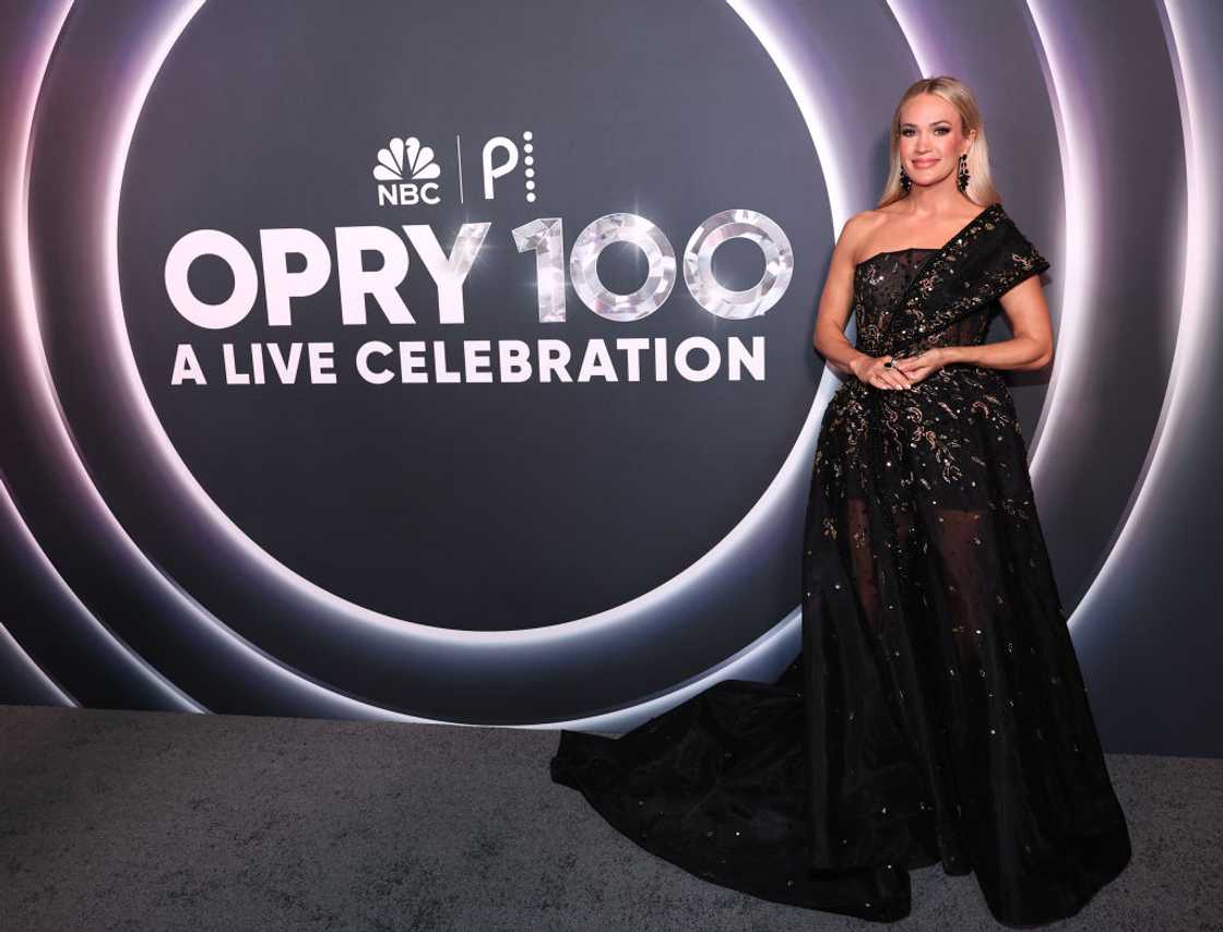 Carrie Underwood with long blonde hair in a black sheer embellished gown, standing in front of a neon Opry 100 backdrop at the Grand Ole Opry in Nashville, 2025 Carrie Underwood with long blonde hair in a black sheer embellished gown, standing in front of a neon Opry 100 backdrop at the Grand Ole Opry in Nashville, 2025